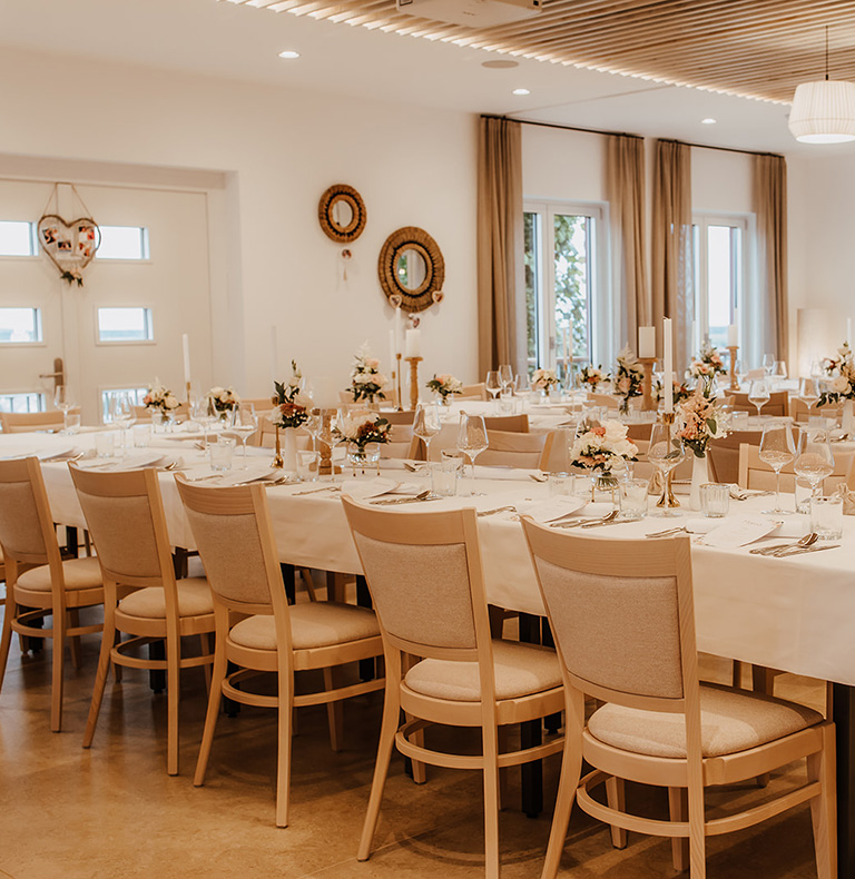 Eleganter Essbereich mit langen, für eine Veranstaltung gedeckten Tischen mit weißen Tischdecken, Blumenschmuck und gedämpfter Beleuchtung, die ein warmes, einladendes Ambiente schaffen.