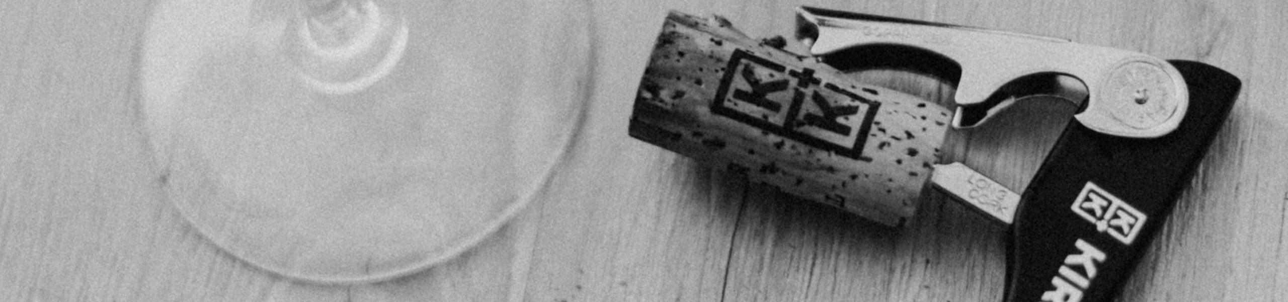 Nahaufnahme eines Weinkorkens und eines Korkenziehers auf einer hellen Holzoberfläche, neben dem Boden eines Weinglases. Das Bild vermittelt eine ruhige, entspannte Atmosphäre.